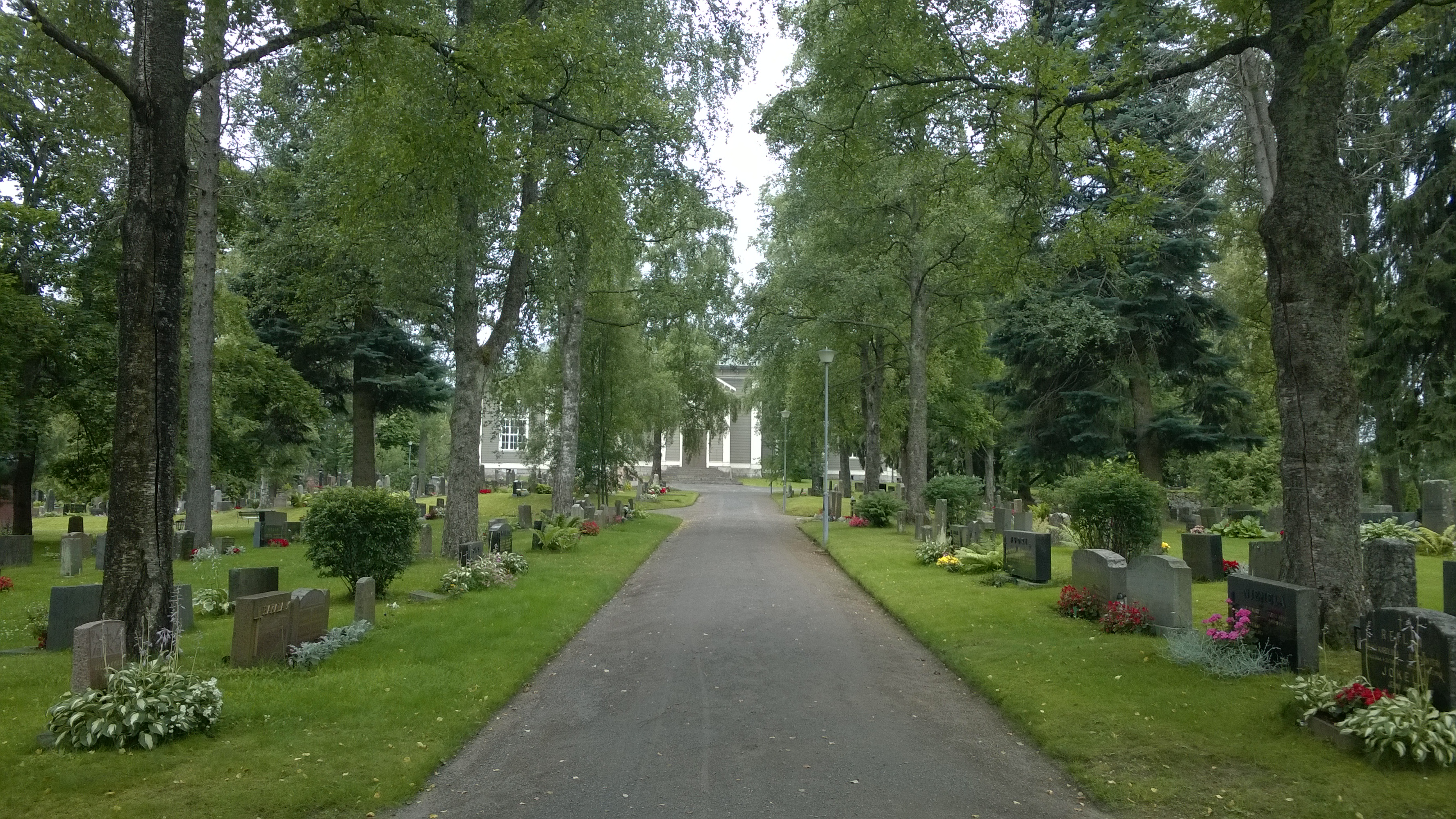 Kesäinen kuva Urjalan hautausmaan rehevien havu- ja lehtipuiden ympäröimältä käytävältä kohti kirkkoa.