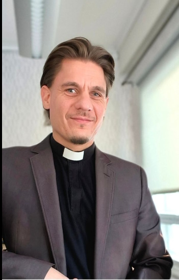 Olli Luhtasela istumassa. Yllään hänellä on harmaa puvuntakki sekä musta papinpaita jossa valkoinen etulipuke.