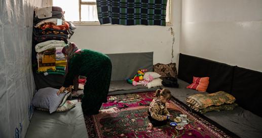 Syyrialaisen perheen äiti lajittelee tekstiilejä ja tyttölapsi leikkii lattialla maton päällä.